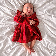 Chloé participe au concours pour gagner de l'argent avec cette photo : baby, infant, newborn, child, red_dress, dress, bed, white_bed, blanket, lying_down, portrait, smiling, hands, feet, cozy, fabric_texture, adorable, indoor, soft_lighting, bedding