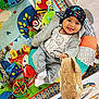 Nikko is registered to the contest to win money with this photo: baby, book, child, clothing, colorful, cute, face, happy, hat, indoors, infant, patterned, playmat, playpen, plush, sitting, smiling, soft_toy, tongue_out, toy