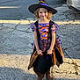 child, costume, witch_hat, purple_dress, black_bucket, smiling, curly_hair, boots, outdoor, driveway, concrete, sunlight, shadow, halloween, trick_or_treat, festive, young_girl, holiday, cute, portrait