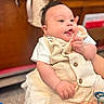 baby, child, infant, cute, playful, tongue_out, beige_outfit, white_shirt, chubby_cheeks, hand, ring, bracelet, indoor, blurred_background, wooden_furniture, person, portrait, smile, holding, closeup