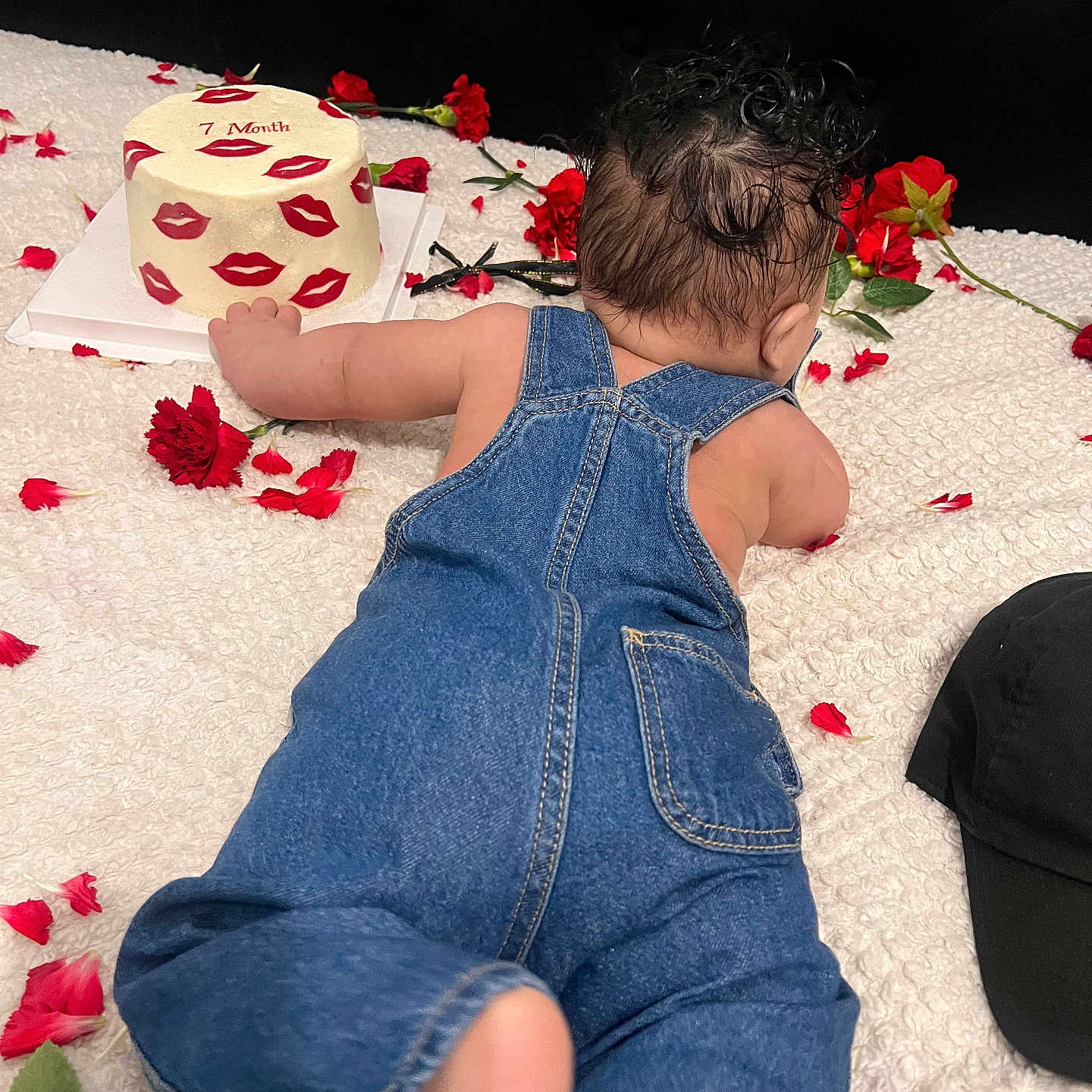 Leyden joined the competition — help win amazing prizes! baby, denim_overalls, cake, red_lips, flower_petals, white_blanket, celebration, infant, back_view, cute, soft_texture, indoor, young_child, skin, hair, crawling, small_cake, red_flowers, black_hat, decor