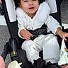Joy is registered to the contest to win money with this photo: toddler, child, stroller, curly_hair, white_outfit, snack_bag, pink_stroller, outdoor, happy, smiling, baby, seatbelt, jacket, footwear, person, cute, infant, holding, daylight, portrait