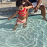 child, toddler, pool, water, splashing, adult, summer, outdoor, sunlight, happy, playing, smiling, curly_hair, flower_accessory, casual_clothing, bracelet, chair, backyard, fence, relaxation