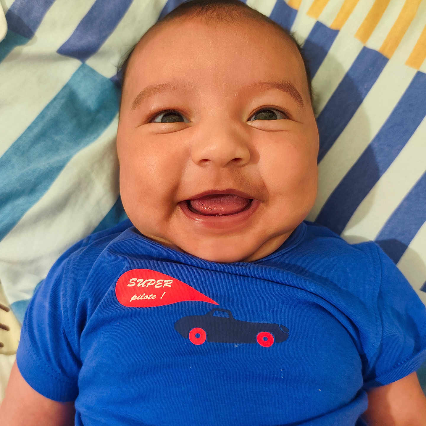 Ethan Paul a rejoint le concours — aidez-le/la à gagner de superbes lots ! baby, bed, blanket, blue_shirt, car_graphic, chubby_cheeks, closeup, clothing, eyes, face, happy, infant, lying_down, mouth, person, portrait, skin, smiling, striped_pattern, tongue