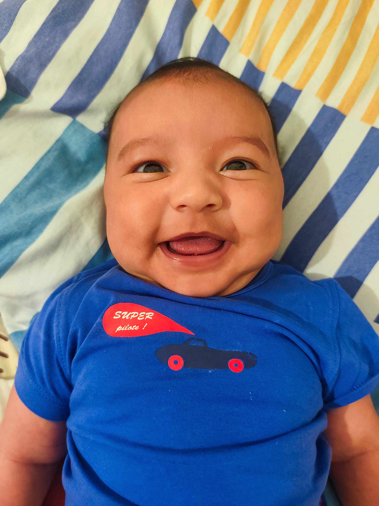 Ethan Paul a rejoint le concours — aidez-le/la à gagner de superbes lots ! baby, infant, smiling, blue_shirt, car_graphic, blanket, striped_pattern, bed, happy, chubby_cheeks, face, eyes, tongue, mouth, lying_down, closeup, skin, person, clothing, portrait