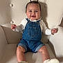 baby, child, denim_overalls, white_shoes, armchair, smiling, happy, cute, indoor, seated, hair_tufts, clothing, infant, face, furniture, portrait, small_hands, legs, skin, cozy