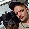 man, dog, rottweiler, cap, selfie, indoor, portrait, pet, animal, face, closeup, headwear, chain_necklace, furniture, railing, wall, expression, casual, companion, friendship