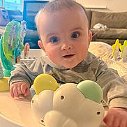 Emmitt is registered to the contest to win money with this photo: baby, infant, face, big_eyes, smile, toy, activity_center, play_table, plastic_toy, hand, sleeve, onesie, indoor, living_room, sofa, couch, pillow, portrait, cute, gaze