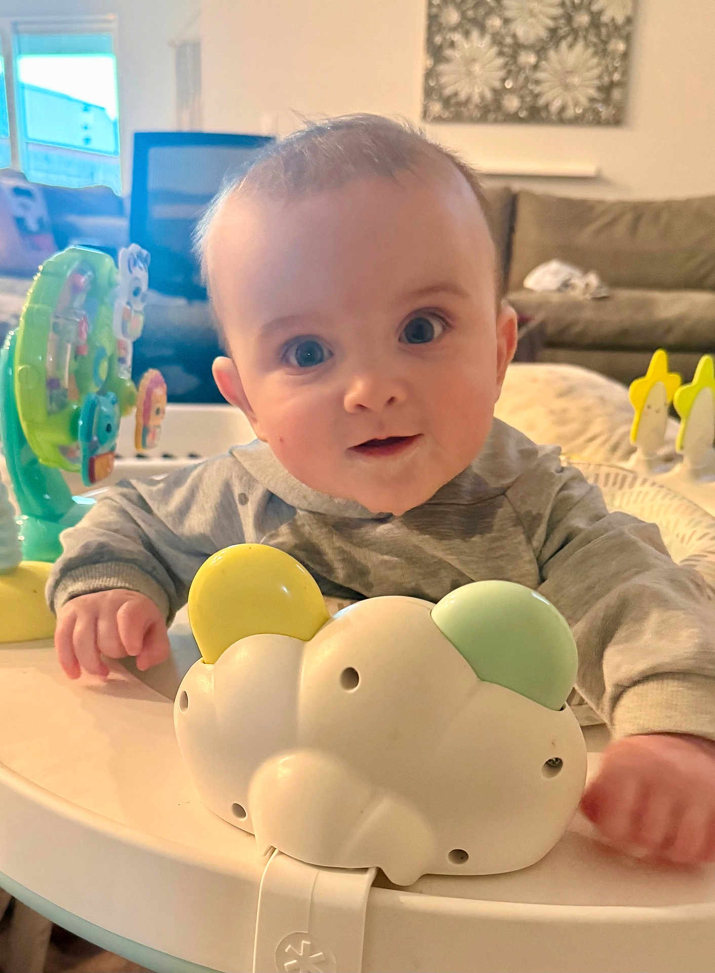 Emmitt is registered to the contest to win money with this photo: baby, infant, face, big_eyes, smile, toy, activity_center, play_table, plastic_toy, hand, sleeve, onesie, indoor, living_room, sofa, couch, pillow, portrait, cute, gaze