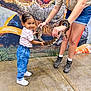 child, girl, snake, reptile, adult, holding, smiling, curious, animal, indoor, concrete_floor, backdrop, casual_clothing, jeans, sneakers, shorts, tshirt, bracelet, watch, interaction