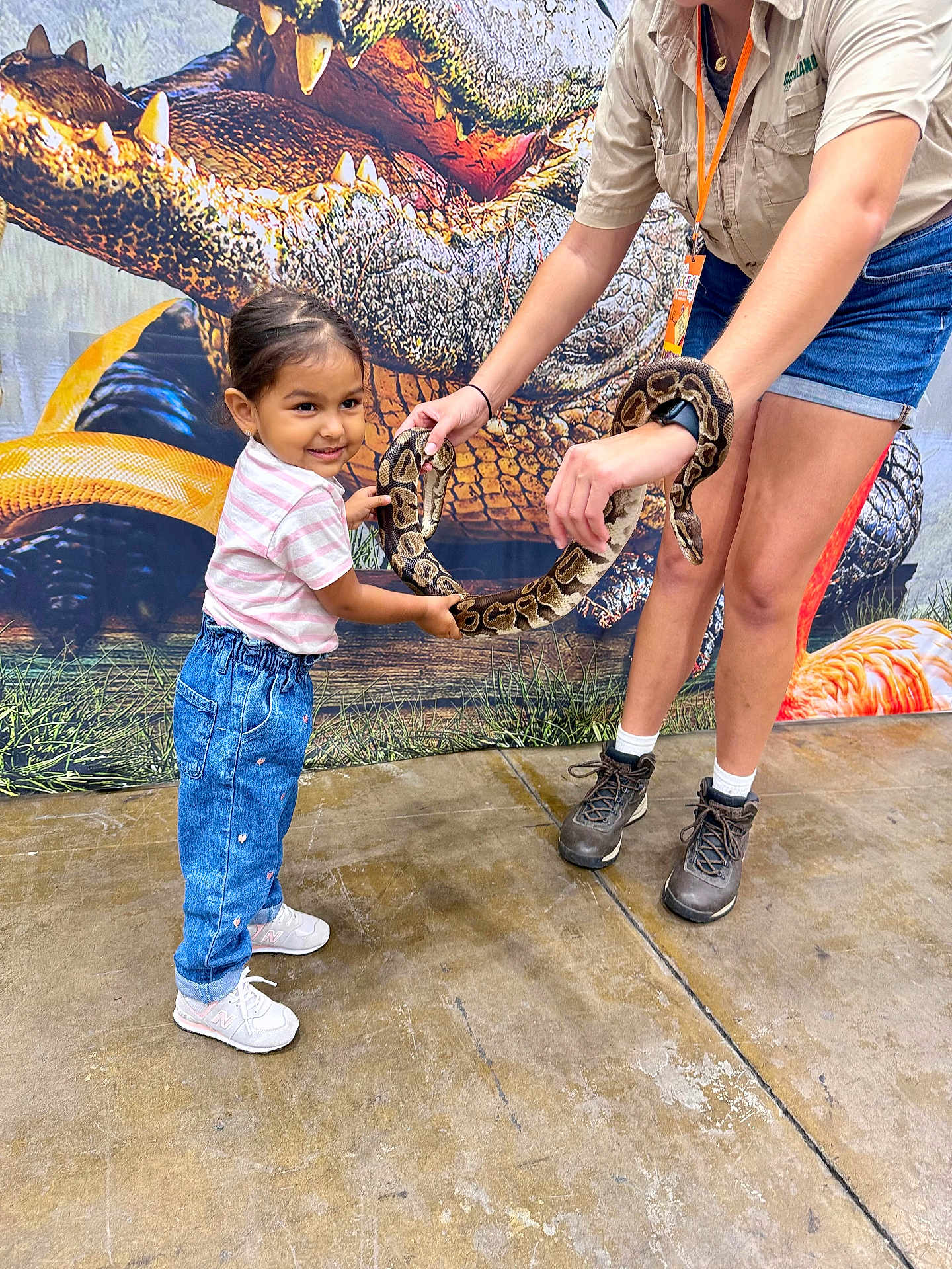 Alayna is registered to the contest to win money with this photo: child, girl, snake, reptile, adult, holding, smiling, curious, animal, indoor, concrete_floor, backdrop, casual_clothing, jeans, sneakers, shorts, tshirt, bracelet, watch, interaction