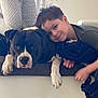 boy, dog, couch, indoor, smiling, resting, friendship, pet, comfort, child, black_and_white_dog, casual_clothing, floor_tiles, blanket, relaxed, portrait, home, human, animal, bond