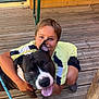 boy, dog, pet, smile, outdoor, wooden_deck, leash, happy, child, black_dog, white_markings, tongue_out, casual_clothing, summer, playing, friendship, crouching, sunlight, wooden_table, green_metal