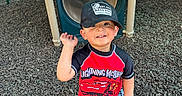David is registered to the contest to win money with this photo: child, toddler, cap, hat, playground, play_structure, wood_chips, smile, waving, outdoor, shorts, shirt, sandals, person, daylight, happy, toy_car_design, casual_clothing, recreation, fun