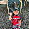 child, toddler, cap, hat, playground, play_structure, wood_chips, smile, waving, outdoor, shorts, shirt, sandals, person, daylight, happy, toy_car_design, casual_clothing, recreation, fun