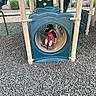 David is registered to the contest to win money with this photo: child, playground, slide, tunnel, cap, spiderman, shorts, outdoor, play_equipment, wood_chips, daylight, person, clothing, sitting, recreation, fun, park, casual, shoes, hat