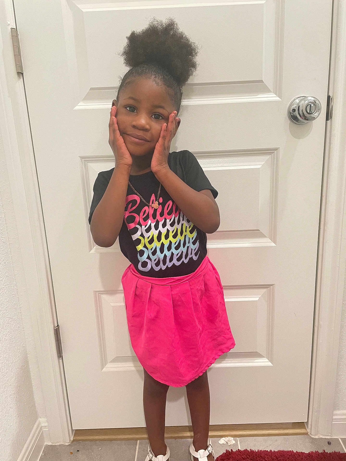 Khalani Cockrell joined the competition — help win amazing prizes! child, girl, standing, indoor, door, black_shirt, pink_skirt, smiling, hands_on_face, hair_puff, white_door, floor_tile, necklace, playful, portrait, casual_clothing, young, cute, happy, person