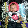 baby, infant, child, stroller, pink_hat, striped_shirt, ladybug_design, jeans, white_shoes, blanket, cushion, zebra_pattern, indoor, cute, young_child, seated, clothing, footwear, headwear, portrait