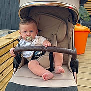 Reggie joined the competition — help win amazing prizes! baby, pacifier, stroller, beige, child, outdoor, wooden_deck, orange_trash_bin, greenery, curious, infant, sitting, clothing, safety_bar, person, face, head, foot, hand, daylight