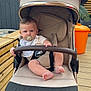 baby, pacifier, stroller, beige, child, outdoor, wooden_deck, orange_trash_bin, greenery, curious, infant, sitting, clothing, safety_bar, person, face, head, foot, hand, daylight