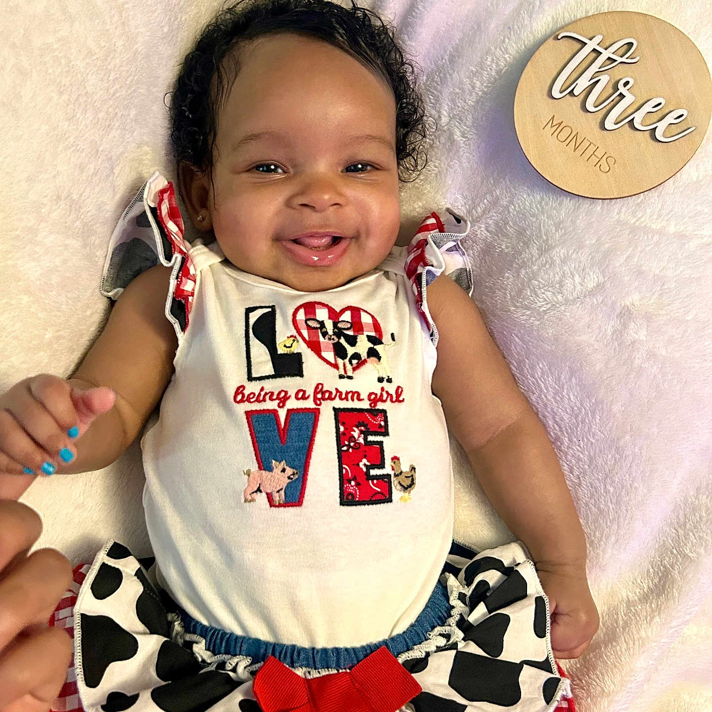 Azanyi is registered to the contest to win money with this photo: baby, child, closeup, curly_hair, cute_outfit, cuteness, farm_girl, hand_holding, happy, indoors, infant, lying_down, portrait, red_bow, skin_tone, skirt, smiling, three_months, white_blanket, wooden_sign