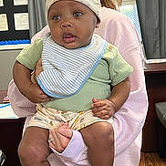 Knoledge is registered to the contest to win money with this photo: baby, child, infant, person, hat, bib, green_shirt, shorts, socks, shoes, hand, adult, indoor, room, furniture, window, curtain, floor, expression, holding