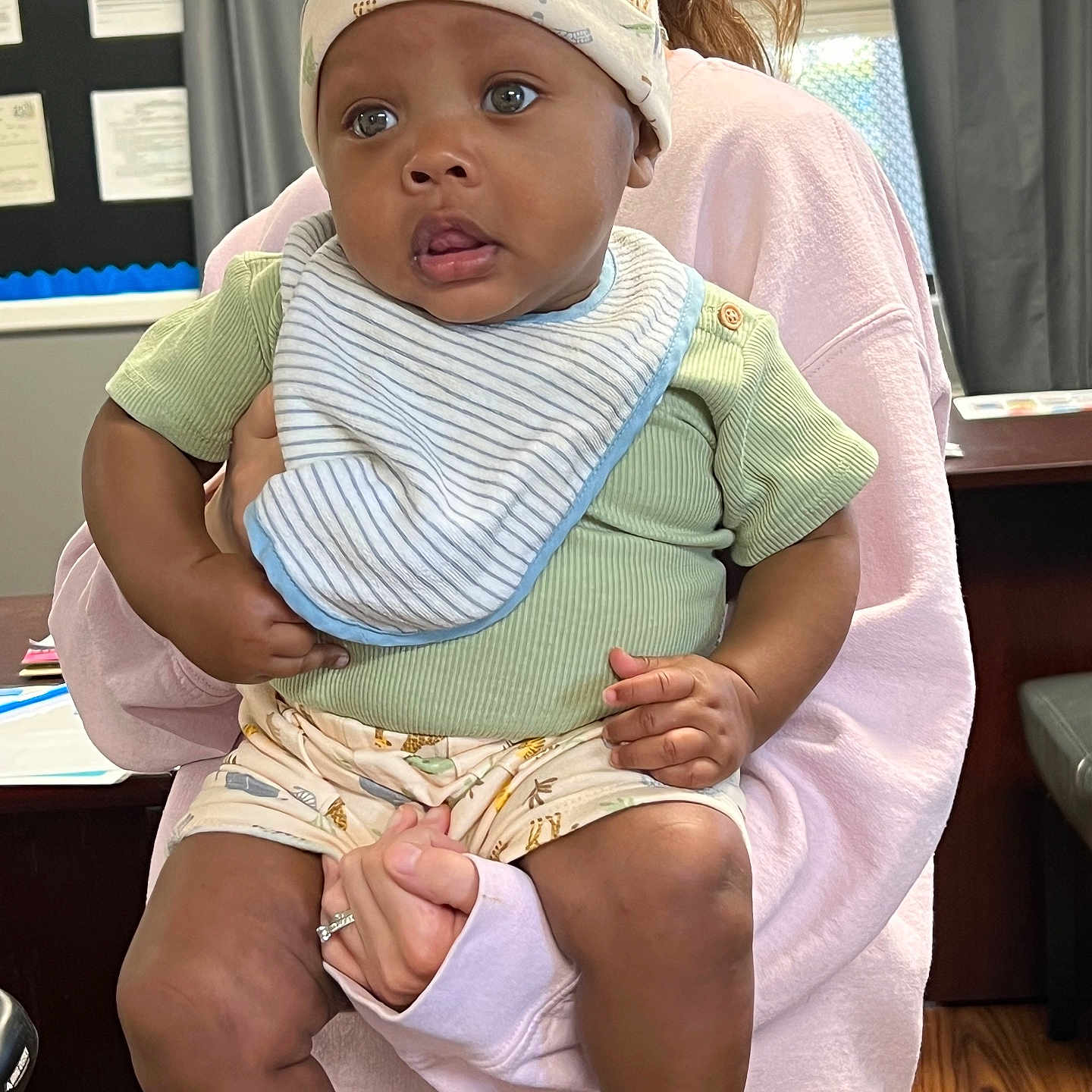 Knoledge is registered to the contest to win money with this photo: adult, baby, bib, child, curtain, expression, floor, furniture, green_shirt, hand, hat, holding, indoor, infant, person, room, shoes, shorts, socks, window