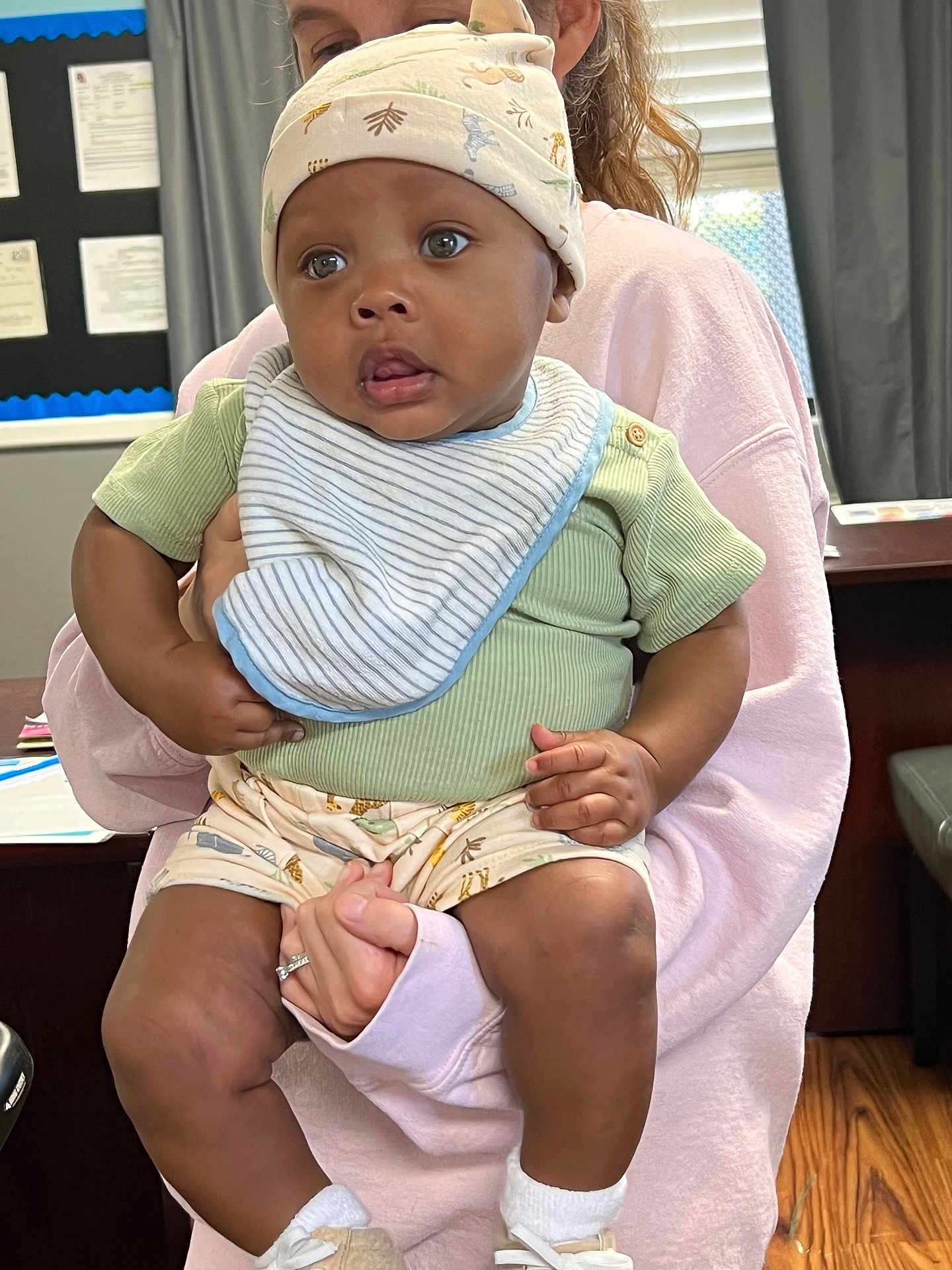 Knoledge is registered to the contest to win money with this photo: baby, child, infant, person, hat, bib, green_shirt, shorts, socks, shoes, hand, adult, indoor, room, furniture, window, curtain, floor, expression, holding