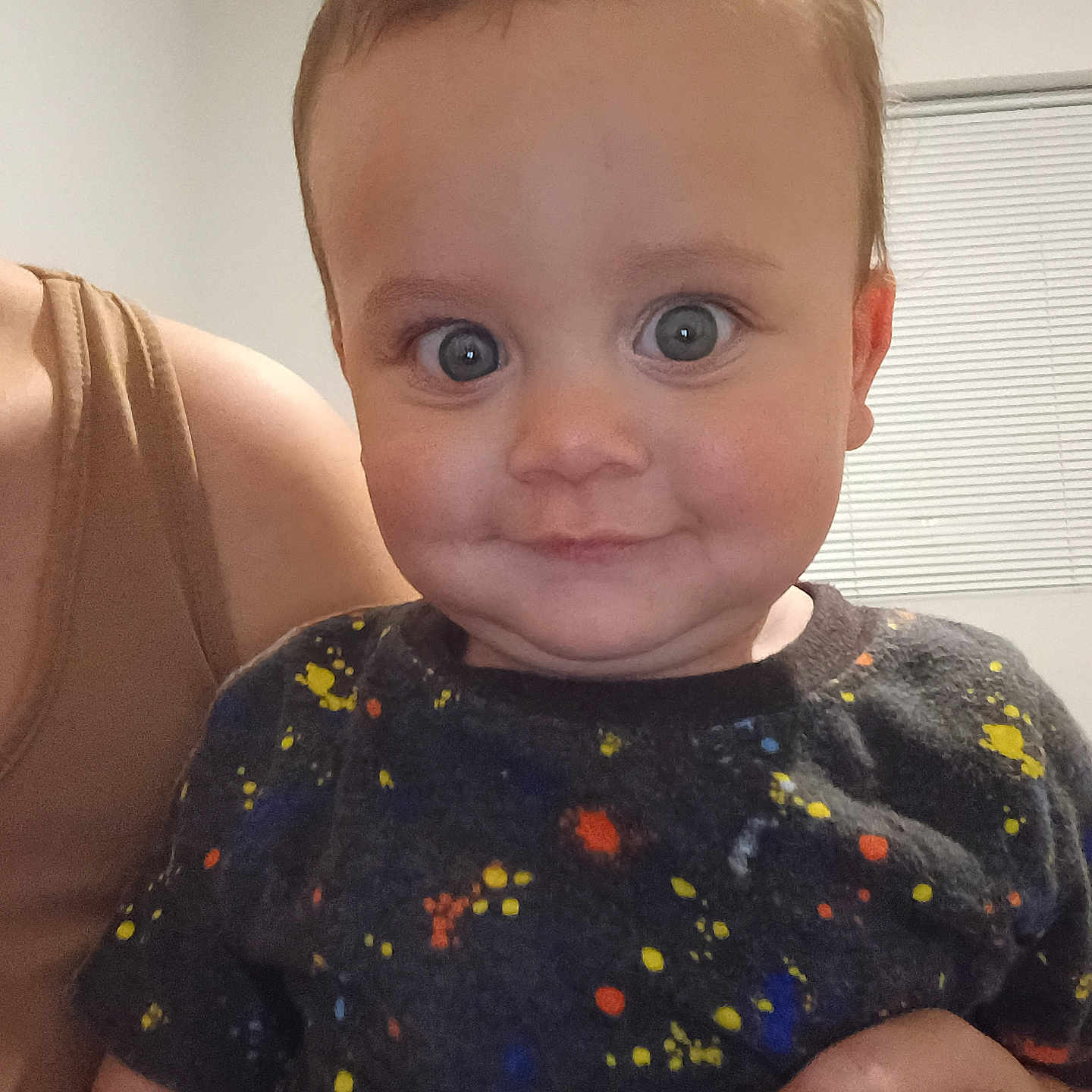 Jimmy is registered to the contest to win money with this photo: baby, child, person, smile, face, indoor, window, blind, adult_hand, colorful_shirt, short_hair, skin, happy, cute, portrait, closeup, holding, expression, eyeballs, casual_clothing