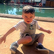 Mathis participe au concours pour gagner de l'argent avec cette photo : toddler, child, boy, sitting, outdoor, sunlight, pool, concrete, overalls, striped_shirt, sneakers, shorts, sunglasses, pocket, arms_outstretched, happy, curious, daytime, casual_clothing, playful