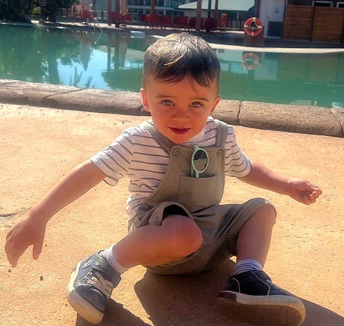 Mathis participe au concours pour gagner de l'argent avec cette photo : toddler, child, boy, sitting, outdoor, sunlight, pool, concrete, overalls, striped_shirt, sneakers, shorts, sunglasses, pocket, arms_outstretched, happy, curious, daytime, casual_clothing, playful