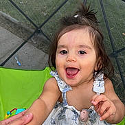 Adeline is registered to the contest to win money with this photo: toddler, child, smiling, hand_holding, floral_dress, outdoor, green_chair, chain_link_fence, grass, person, happy, cute, playful, young_child, summer, daylight, toy, baby, portrait, casual_clothing