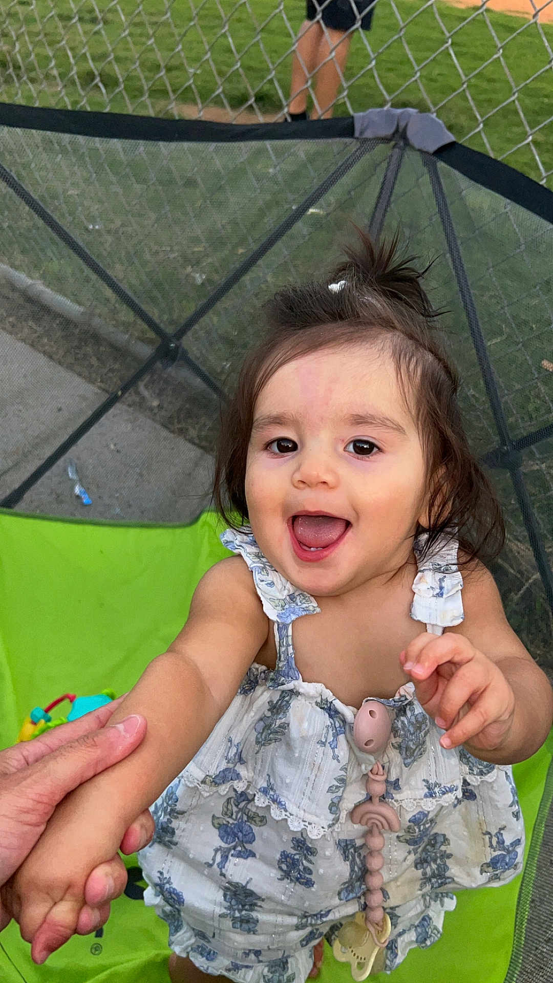 Adeline is registered to the contest to win money with this photo: toddler, child, smiling, hand_holding, floral_dress, outdoor, green_chair, chain_link_fence, grass, person, happy, cute, playful, young_child, summer, daylight, toy, baby, portrait, casual_clothing