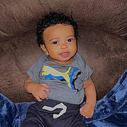 Amir is registered to the contest to win money with this photo: baby, child, curly_hair, gray_shirt, pants, socks, blue_blanket, brown_chair, smiling, indoor, comfortable, cute, young_child, portrait, seated, happy, playful, soft_texture, casual_clothing, human