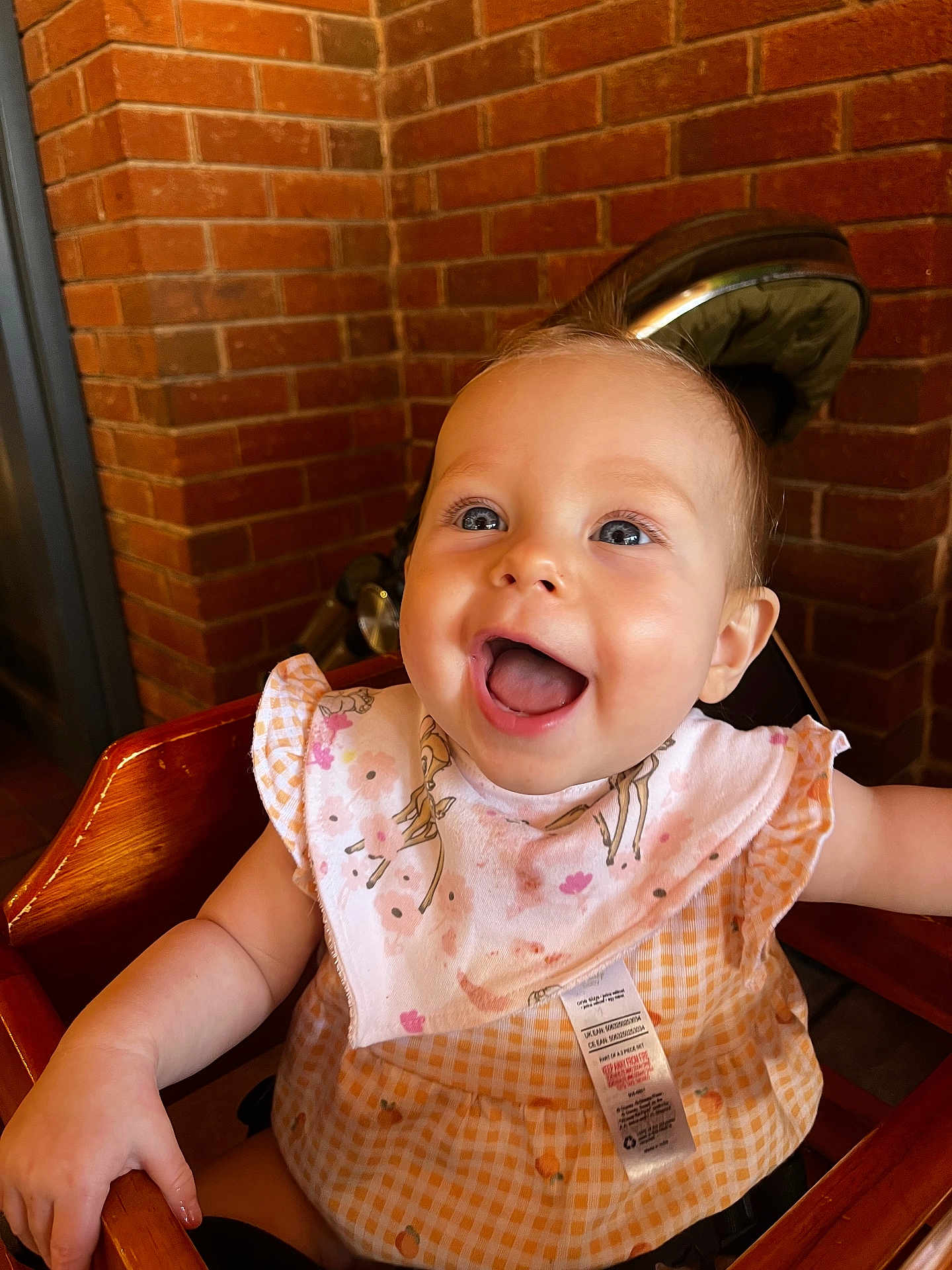 Amélie is registered to the contest to win money with this photo: baby, child, high_chair, smiling, happy, indoor, bib, clothing, face, person, wood, furniture, gingham, pattern, pink, orange, deer, floral, brick_wall, cute