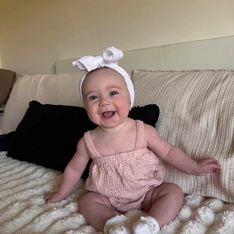 Lucy joined the competition — help win amazing prizes! baby, infant, child, smiling, headband, pink_outfit, sitting, blanket, couch, pillow, indoors, happy, cute, portrait, young_child, soft_texture, cozy, person, toddler, head