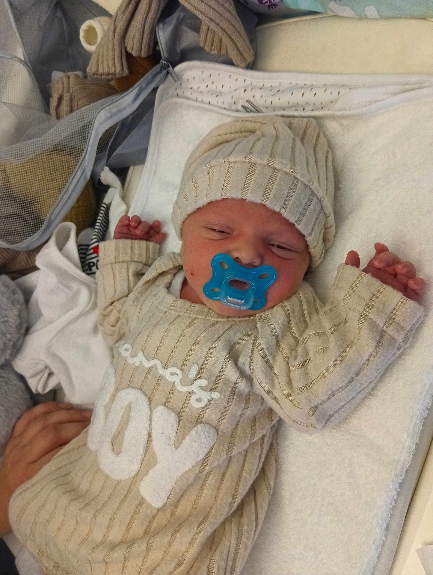 Mathëo participe au concours pour gagner de l'argent avec cette photo : newborn, baby, pacifier, blue_pacifier, ribbed_hat, striped_onesie, joy_text_on_clothing, blanket, towel, bassinet, hospital_setting, sleeping, hands, adult_hand, soft_fabric, portrait, cozy, resting, infant_clothing, nursery_items