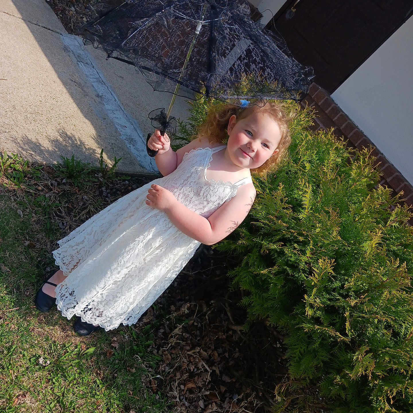 Aurora is registered to the contest to win money with this photo: black_umbrella, bush, child, curly_hair, dress, footwear, girl, grass, greenery, happy, nature, outdoor, person, playful, portrait, sidewalk, smile, sunlight, umbrella, white_dress