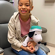 Raelynn is registered to the contest to win money with this photo: child, girl, smiling, plush_toy, snoopy, jeans, hoodie, medical_office, chair, happy, indoor, casual_clothing, person, seated, toy, healthcare, cute, young, relaxed, waiting_room