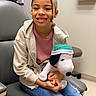 child, girl, smiling, plush_toy, snoopy, jeans, hoodie, medical_office, chair, happy, indoor, casual_clothing, person, seated, toy, healthcare, cute, young, relaxed, waiting_room
