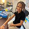child, girl, smiling, braided_hair, bracelet, classroom, table, bookshelf, chair, pillow, educational_posters, school, black_polo, khaki_skirt, happy, seated, indoor, learning, young, cute