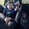 child, toddler, car_seat, toy, cow_toy, smiling, happy, car_interior, seatbelt, backseat, window, daylight, blue_shirt, shorts, backpack, child_safety, portrait, cute, playful, young_child