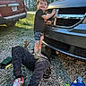 child, adult, car, vehicle, outdoor, gravel, smile, person, automobile, trees, daylight, repair, helping, license_plate, shoe, pants, tshirt, metal, nature, grille