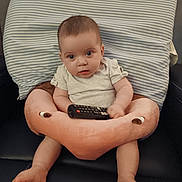 Abigail Joy is registered to the contest to win money with this photo: baby, infant, remote_control, plush_cushion, flamingo_cushion, striped_pillow, chair, seated, indoor, clothing, short_sleeves, curious, looking_at_camera, person, child, skin, legs, feet, face, expression