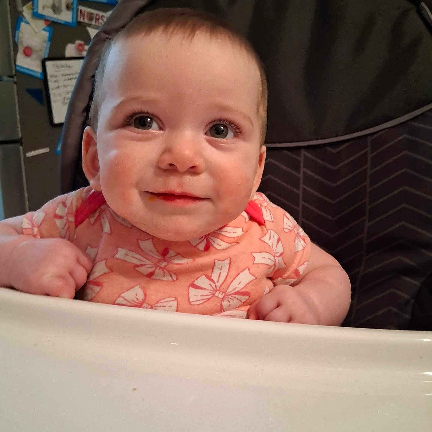 Abigail Joy is registered to the contest to win money with this photo: baby, child, high_chair, pink_clothing, floral_pattern, smiling, face, eyes, kitchen, fridge, magnets, notes, curly_hair, cute, infant, indoor, person, happy, table, seat
