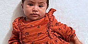 Nylah is registered to the contest to win money with this photo: baby, infant, child, dress, orange_clothing, floral_pattern, lying_down, white_background, cute, portrait, face, hair, hand, fist, young_child, skin, person, indoors, soft_texture, serious_expression