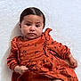 baby, infant, child, dress, orange_clothing, floral_pattern, lying_down, white_background, cute, portrait, face, hair, hand, fist, young_child, skin, person, indoors, soft_texture, serious_expression