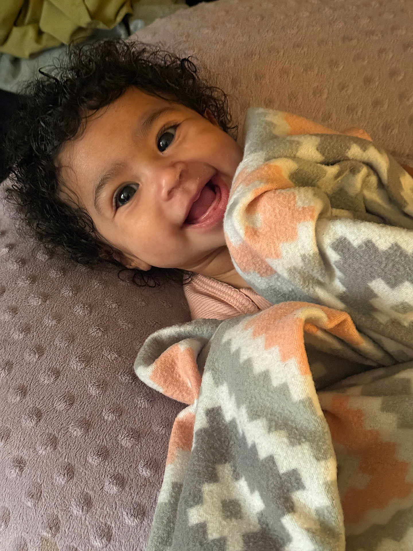 Nylah is registered to the contest to win money with this photo: baby, smile, curly_hair, blanket, patterned_blanket, infant, happy, lying_down, soft_texture, face, child, cozy, indoors, wrapped, cute, expression, person, warm, comfort, toddler