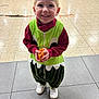 Charly a rejoint le concours — aidez-le/la à gagner de superbes lots ! child, toddler, smiling, costume, green, red, white_shoes, indoor, floor_tiles, chairs, orange_wrapped_treat, happy, cute, person, standing, playful, clothing, young_child, portrait, casual