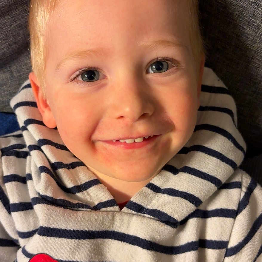 Charly a rejoint le concours — aidez-le/la à gagner de superbes lots ! blue_eyes, casual_clothing, child, close_up, couch, cute, fabric, face, happy, human, indoors, kid, person, playful, portrait, smiling, striped_hoodie, toy_vehicle, yellow_toy, young_child